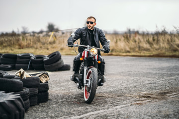 Bearded man in sunglasses and leather jacket sitting on a red motorcycle and looking on the road. Sideways along the road lie tires