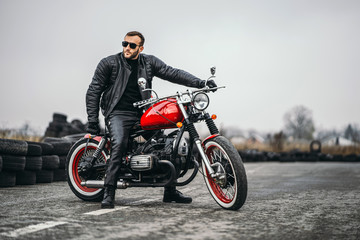 Red motorbike with rider. A man in a black leather jacket and pants stands sideways in the middle of the road. Tires are laid on the background