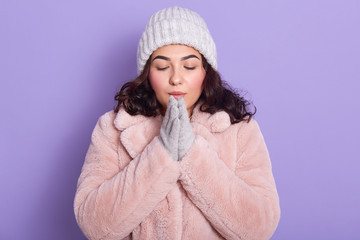 Beautiful young woman basking her hands in winter, wearing warm clothes, faux fur coat, gloves and cap, posing isolated over lilac background, stands with closed eyes. Winter and cold weather concept.
