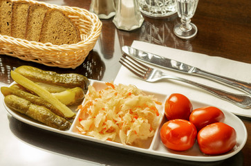 White dish with canned cucumbers, cabbage and tomatoes, with bread and shot glass with a drink on the table.
