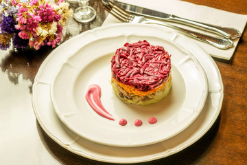 White plate on the brown wooden table in restaurant with salad herring with vegetables: potato, carrot and beet.