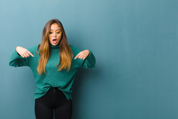 young pretty woman feeling shocked, open-mouthed and amazed, looking and pointing downwards in...