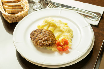 Fried cutlet with mashed potatoes and red fresh tomato on the white plate on the brown table. Serving of restaurant.