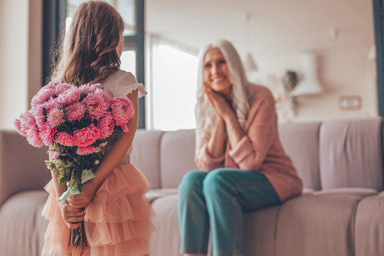 girl holding bouquet of flowers in the hands behind her back for granny sitting on the couch