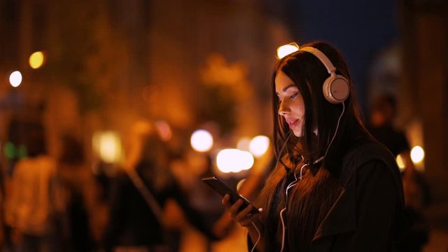 Beautiful young woman in headphones and with mobile phone in the night city. Stylish girl woman texting and listening to music on phone in the city at night