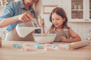 grandmother pouring milk from the jug into the bowl with flour for cookie dough and granddaughter...