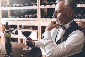 Sommelier Concept. Senior man sitting at table stirring wine in glass concerned side view close-up