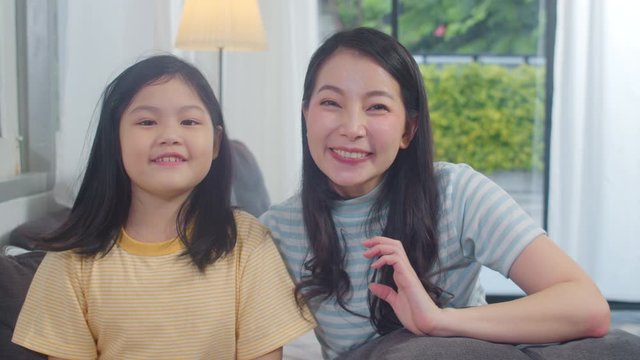 Asian woman and daughter video call at home. Mother and kid happy spend time together using mobile phone video call talking with dad on sofa in living room at modern home in the evening concept.