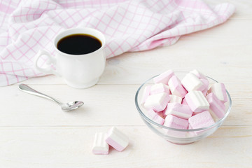 Obraz na płótnie Canvas White pink fluffy marshmallows in a glass bowl and a cup of coffee on a light wooden table. Sweet and tasty winter food. Delicious soft sweets.
