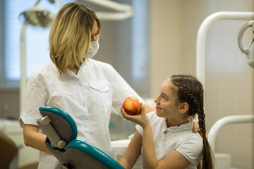 Female dentist gives apple for smiling teenage girl in modern dental clinic.