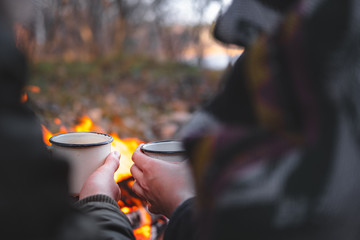 Two people warming hands with hot drinks by the bonfire. Spending nice time outdoors in chilly weather at a camping place - tranquil and peaceful scene