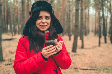 A portrait of the pretty woman with a cup of tea in woods. The woman in a jacket and a hat with earflaps is drinking a tea in a cold weather.