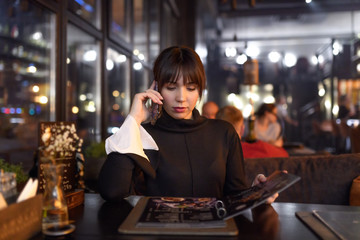 Brunette Woman in black shirt talking on cellphone while making a choice of a dish or drink in cafe. Discussing order with girlfriend by phone.