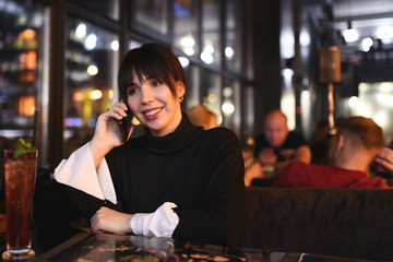 cheerful and smiling Woman in black shirt talking on cellphone while waiting her partner and lover in cafe. Discussing order with girlfriend by phone.