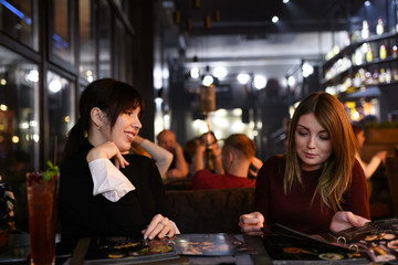 Two girlfriends in hookah shisha lounge bar making an order. One is choosing dish and second is waiting until she make a decision.