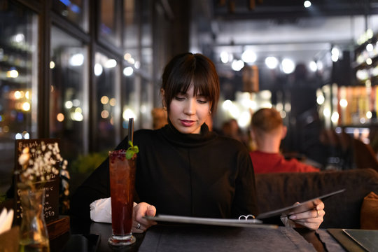 Beautiful Young Caucasian Woman Sitting In Restaurant And Attentively Looking At Menu Choosing What To Order. Dinner Outdoor. Visiting Cafe In Evening After Job.