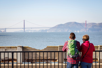 San Francisco, California, USA: A tourist couple enjoying the view over the bay with the Golden Gate Bridge from the Aquatic Park Pier.