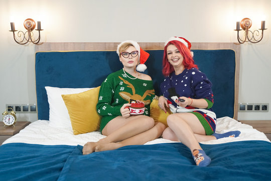 lifestyle indoor portrait of two young woman posing in Christmas sweaters on the bedroom at New Year eve. Smiling having fun, ready for celebration. Bright holiday image of best friends.