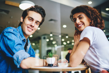 Below portrait of positive couple in love smiling at camera sitting at coffee table in modern cafeteria.Happy young girlfriend and boyfriend in casual wear spending leisure time together