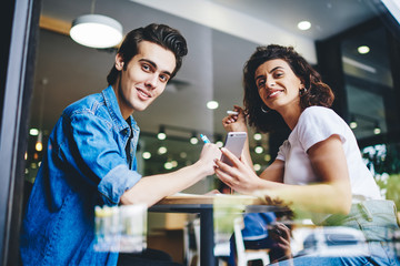 Portrait of cheerful young man and woman dressed in casual wear smiling at camera while sitting together in cafe and installing mobile app on cellular.Happy couple in love spending time in coffee shop