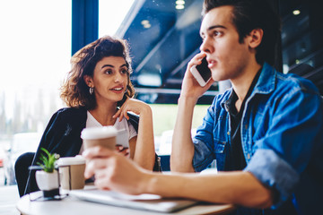 Young man calling to operator on smartphone about setting of mobile benefits of roaming connection while spending leisure time together with stylish hipster girl in modern coffee shop