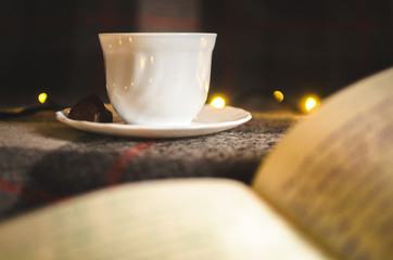 A cup of hot black tea with chocolate on a saucer on a warm and cozy plaid with Christmas lights