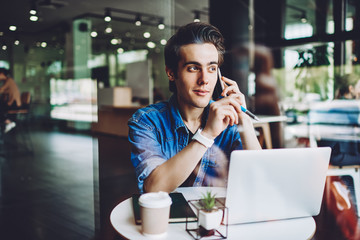 Casual dressed hipster guy talking on smartphone for discussing important information sitting with laptop in coworking space, young man calling on cellular to customer service for consultancy