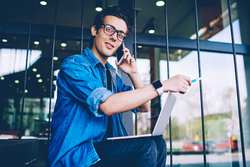 Portrait of positive hipster guy in eyeglasses spending time in campus and looking at camera during calling to customer service for communicated about software updating on new laptop computer