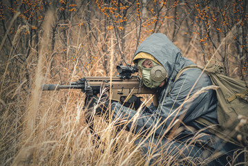 Soldier with airsoft rifle in raincoat and gas mask outdoor.