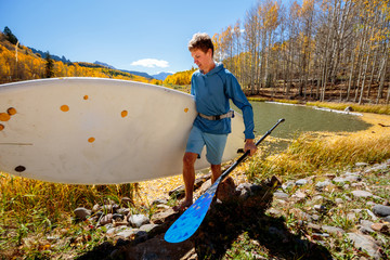 A man carries a SUP board away froma an alpine lake near Telluride, Colorado in autumn in the San Juan Mountains.