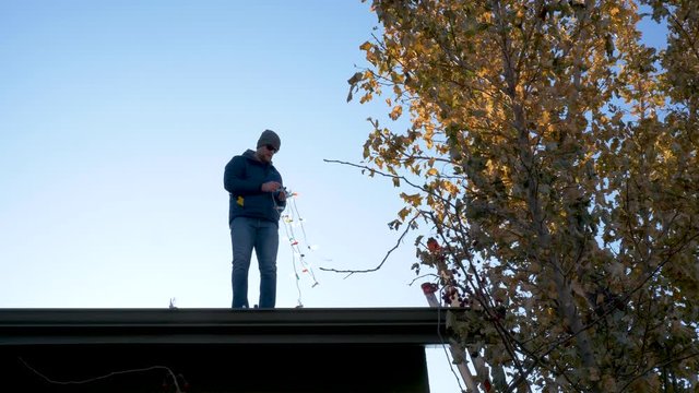 Man standing on the roof of his home untangles a strand of Christmas lights