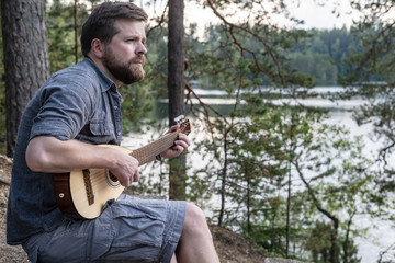 Lonely bearded man plays a melody on a guitar and is sad, sitting on a hill, against a background of trees and a lake.