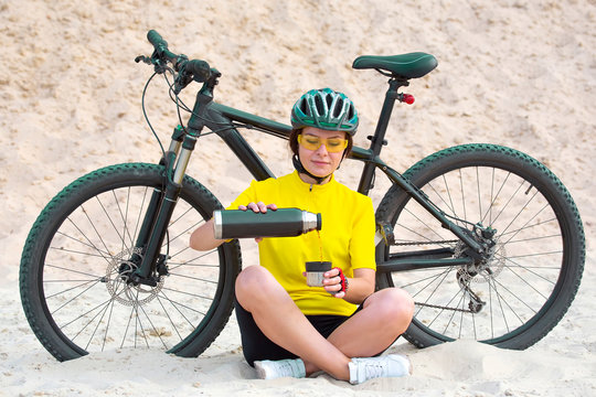 Beautiful girl cyclist pours tea from a thermos on the background of a Bicycle in the sand. Sports and recreation.
