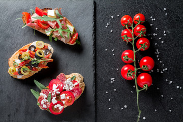 Traditional Bruschetta. Italian antipasti set with jamon, guanchial sausage, olives, cottage cheese, arugula and tomatoes on a black background