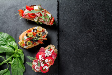 Traditional Bruschetta. Italian antipasti set with jamon, guanchial sausage, olives, cottage cheese, arugula and tomatoes on a black background