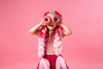 cute little girl in a pink dress with pink braids from kanekalon holds donuts in pink glaze in her...