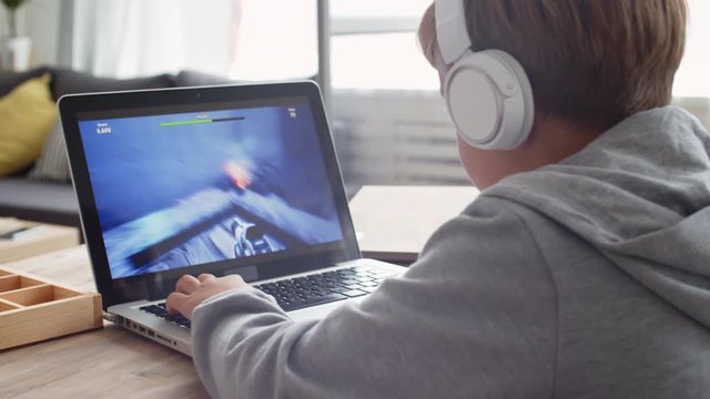 Back view of Caucasian boy wearing grey hoody and wireless headphones sitting at computer and playing computer game