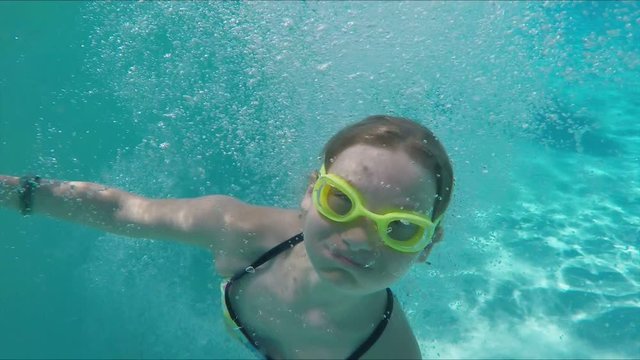 Funny girl in bright swimming goggles dives underwater into the pool and spins around the camera
