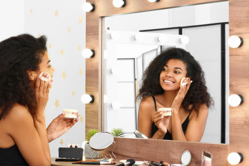 Beautiful African-American woman applying makeup in front of mirror