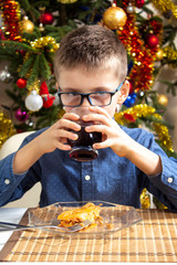 Chłopiec siedzi przy stole podczas obiadu. Trzyma w dłoniach szklankę z napojem i pije z niej. Bożonarodzeniowa choinka w tle.