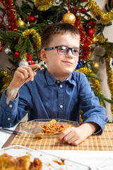 Uśmiechnięty chłopiec podczas Świąt Bożego Narodzenia siedzi przy stole i zjada świąteczne danie. - obrazy, fototapety, plakaty