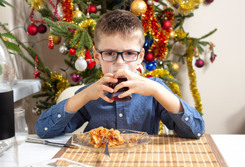 Chłopiec siedzi przy stole podczas obiadu. Trzyma w dłoniach szklankę z napojem i pije z niej. Bożonarodzeniowa choinka w tle. - obrazy, fototapety, plakaty