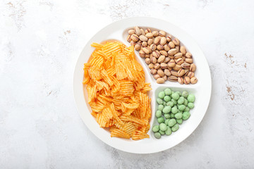 Plate with tasty potato chips, pistachios and peanuts on light background