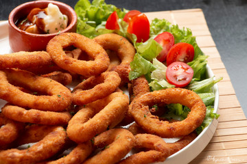 Porção frita de anel de cebolas em prato de louça branco e decoração de tomate e alface, molho