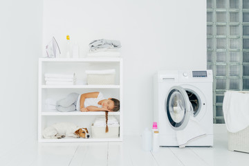 Cozy washing room with washer, sleeping girl with dog on shelf, bottles of liquid powder on floor, basket full of dirty laundry. Child has rest after helping mum to wash clothes. Domestic atmosphere