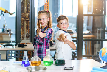 Portrait shot of two cheerful Caucasian scientist kids in safety goggles showing thumbs up in primary school chemical class. Science, chemistry and children. Slow motion