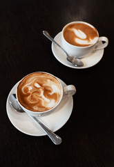Two cups of cappuccino on a textured dark wood table in a cafe with classical interior. Close-up.
