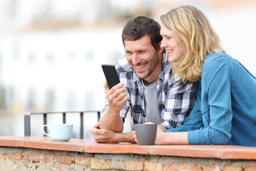 Happy adult couple checking smart phone in a balcony