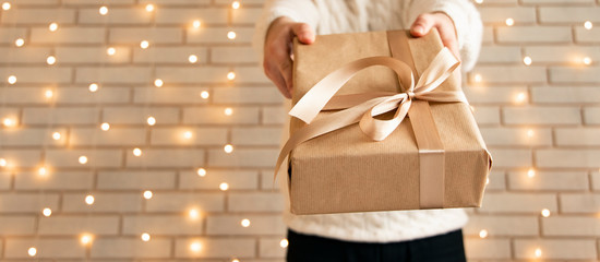 advertising pattern for Christmas holidays or another event congratulation hands taking gift to camera in studio interior white brick wall and yellow garland illumination unfocused background 
