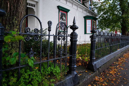 A Large White House With Green And Red Trim On The Windows.The Front Entrance Has A Wrought Iron Fence With Greenery, Trees And Shrubs. There Are Orange Dead Leaves Along The Sidewalk In Front.